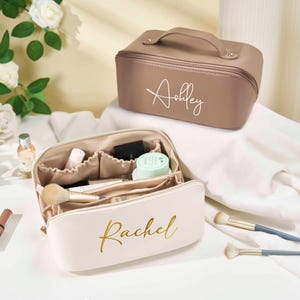 Puede incluir: Dos neceseres de maquillaje personalizados, uno color topo y otro blanco roto, con letras doradas. El neceser color topo tiene un asa y el nombre "Ashley" en cursiva. El neceser blanco roto, etiquetado "Rachel", está abierto, revelando brochas y productos de maquillaje. Un tubo de maquillaje es visible.