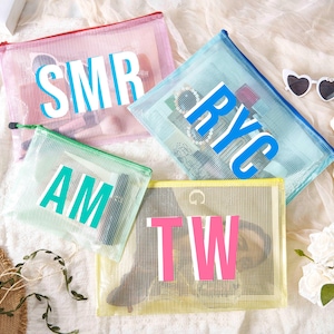 Op de afbeelding: Vier mesh ritszakjes in roze, blauw, groen en geel, elk met witte en turquoise of roze blokletters. De zakjes zijn gevuld met diverse items. Een hartvormige zonnebril en bloemen zijn ook aanwezig.