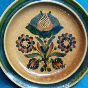 May include: A ceramic plate with a floral design. The plate features a tan center with a blue and brown floral pattern. The rim has a green and blue stripe. The plate is round.