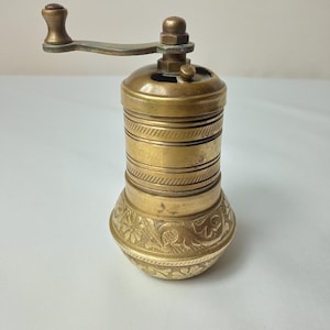 May include: Antique brass pepper mill with a hand crank. The mill has a cylindrical body with horizontal ridges and a floral pattern on the base. The crank and handle are made of brass. The pepper mill is a kitchen utensil.