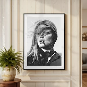 Puede incluir: Impresi&oacute;n enmarcada en blanco y negro de una mujer con el pelo al viento, fumando un cigarrillo. El retrato presenta una bufanda y una chaqueta negras. La imagen se muestra en una pared de color claro con un helecho en una maceta decorativa.