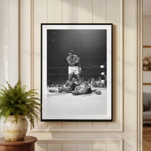 May include: Black and white framed photograph of a boxing match. One boxer stands over another who is lying on the mat. The standing boxer is wearing white shorts and the other boxer is wearing black shorts.