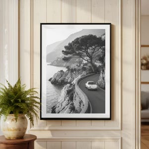 May include: Monochrome framed print showing a white car on a coastal road. The road curves along the coastline, with a stone wall on one side and the sea on the other. A large tree is on the side of the road.