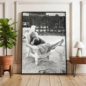 Puede incluir: Impresi&oacute;n enmarcada en blanco y negro de una mujer relaj&aacute;ndose en una silla de madera al aire libre. Viste un top oscuro y una falda a cuadros. La imagen es una fotograf&iacute;a vintage, con un marco negro.