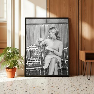 May include: Black and white framed print featuring a woman seated in a chair, holding a purse and a glass, with a cigarette in her mouth. The print is displayed in a room with a potted plant and a wooden side table.