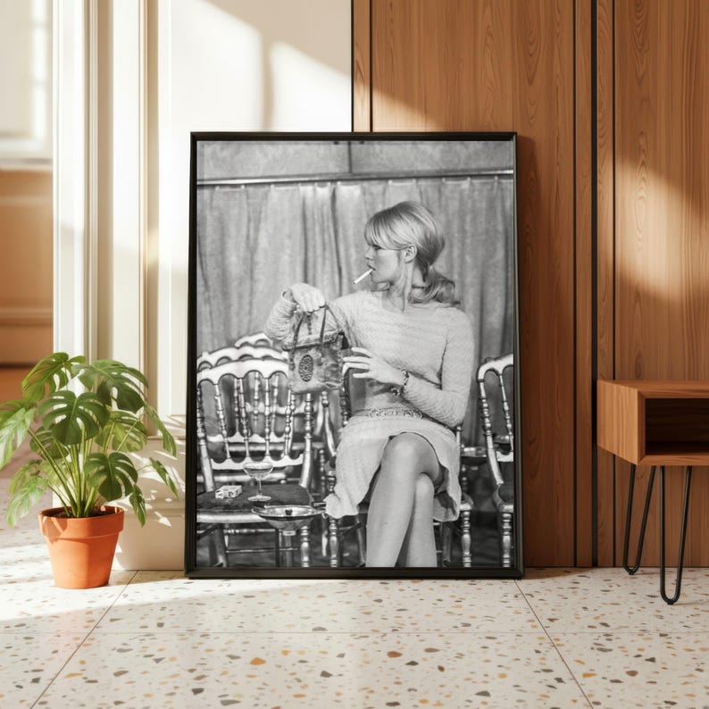 May include: Black and white framed print featuring a woman seated in a chair, holding a purse and a glass, with a cigarette in her mouth. The print is displayed in a room with a potted plant and a wooden side table.