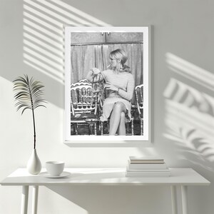 May include: Black and white framed print of a woman seated in a chair, holding a cigarette and a purse. The print is displayed on a white table with a vase, a cup and saucer, and a stack of books. The image is in a white frame.