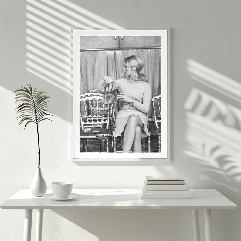 May include: Black and white framed print of a woman seated in a chair, holding a cigarette and a purse. The print is displayed on a white table with a vase, a cup and saucer, and a stack of books. The image is in a white frame.
