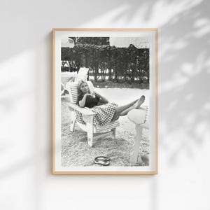 Puede incluir: Fotograf&iacute;a en blanco y negro enmarcada de una mujer relaj&aacute;ndose en una silla de madera. Viste un top oscuro y una falda a cuadros, con sandalias en el suelo. El fondo presenta &aacute;rboles y otras sillas.