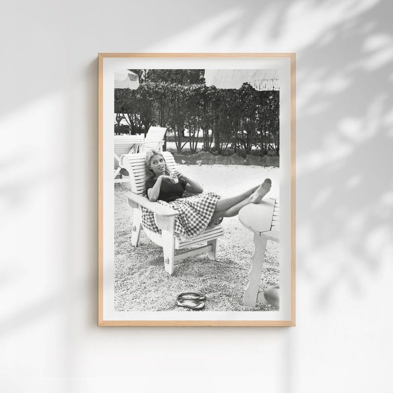 Puede incluir: Fotograf&iacute;a en blanco y negro enmarcada de una mujer relaj&aacute;ndose en una silla de madera. Viste un top oscuro y una falda a cuadros, con sandalias en el suelo. El fondo presenta &aacute;rboles y otras sillas.