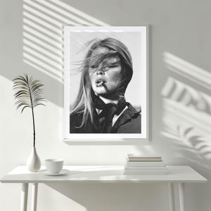 Puede incluir: Impresi&oacute;n en blanco y negro enmarcada de una mujer con el pelo al viento, fumando un cigarrillo. Lleva una bufanda oscura y una chaqueta. La impresi&oacute;n est&aacute; sobre una mesa blanca con un jarr&oacute;n, una taza y una pila de libros.