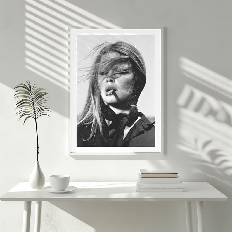 Puede incluir: Impresi&oacute;n en blanco y negro enmarcada de una mujer con el pelo al viento, fumando un cigarrillo. Lleva una bufanda oscura y una chaqueta. La impresi&oacute;n est&aacute; sobre una mesa blanca con un jarr&oacute;n, una taza y una pila de libros.