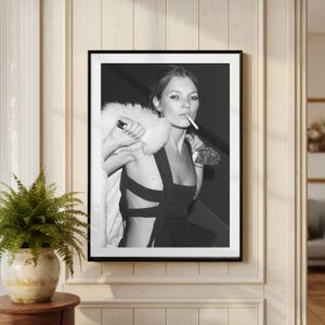 May include: Black and white framed print of a woman smoking a cigarette. She is wearing a black dress and a white fur stole. The image is displayed on a cream-colored wall.