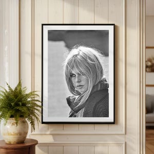 Puede incluir: Retrato en blanco y negro enmarcado de una mujer con cabello rubio y una bufanda. La fotograf&iacute;a se muestra en una pared de color claro, con un helecho en una maceta decorativa en primer plano.