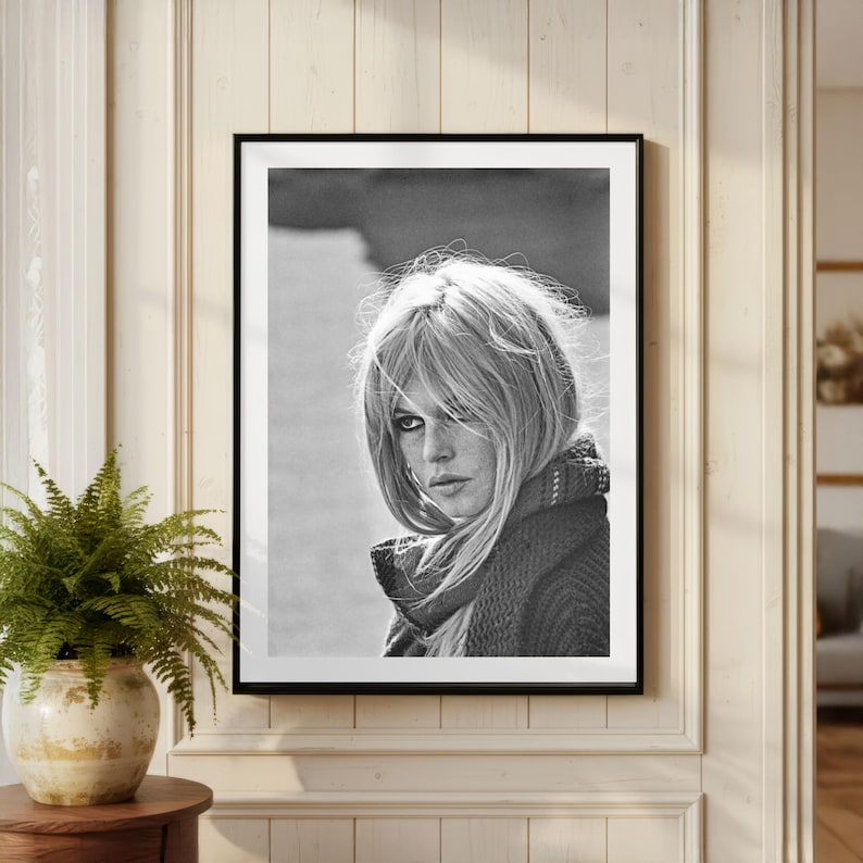 Puede incluir: Retrato en blanco y negro enmarcado de una mujer con cabello rubio y una bufanda. La fotograf&iacute;a se muestra en una pared de color claro, con un helecho en una maceta decorativa en primer plano.