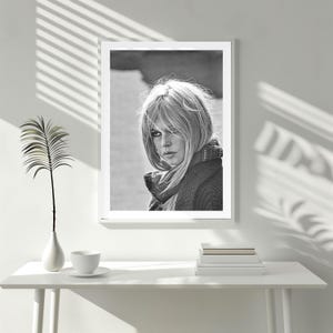 Puede incluir: Retrato enmarcado en blanco y negro de una mujer con cabello rubio, que lleva una bufanda y un su&eacute;ter. La imagen se muestra sobre una mesa blanca con un jarr&oacute;n, una taza y un platillo, y una pila de libros.