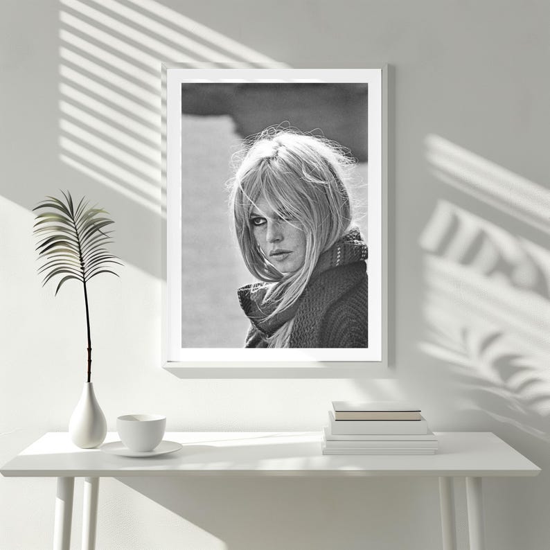 Puede incluir: Retrato enmarcado en blanco y negro de una mujer con cabello rubio, que lleva una bufanda y un su&eacute;ter. La imagen se muestra sobre una mesa blanca con un jarr&oacute;n, una taza y un platillo, y una pila de libros.