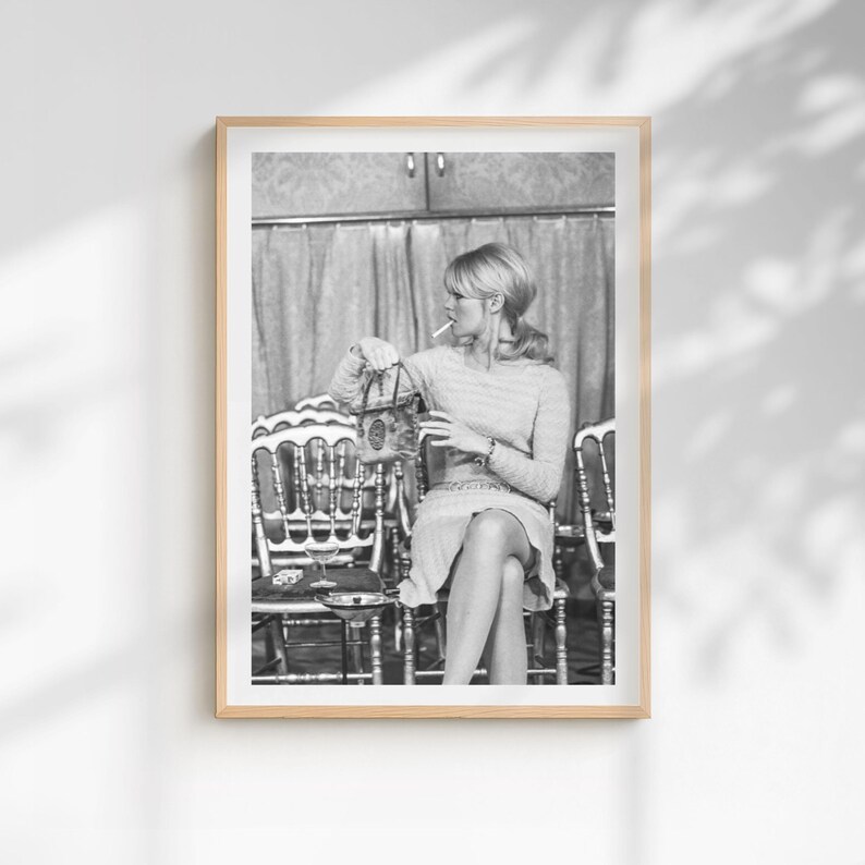 May include: Black and white framed print of a woman seated, holding a purse and smoking a cigarette. She is wearing a light-colored dress and is seated in front of a patterned curtain. The print is in a wooden frame.