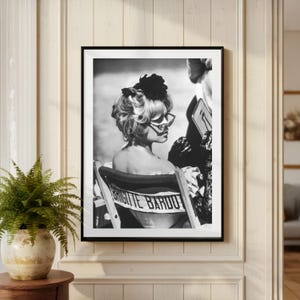 May include: Black and white framed print of a woman in sunglasses and a floral headpiece, seated in a beach chair. The chair has the name "Brigitte Bardot" printed on it. The image is displayed on a white wall.