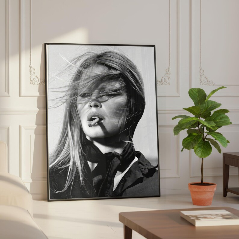 Puede incluir: Impresi&oacute;n enmarcada en blanco y negro de una mujer con cabello largo y un cigarrillo en la boca. Lleva una parte superior oscura y una bufanda. La impresi&oacute;n est&aacute; apoyada contra una pared blanca en una habitaci&oacute;n con una planta y una mesa.