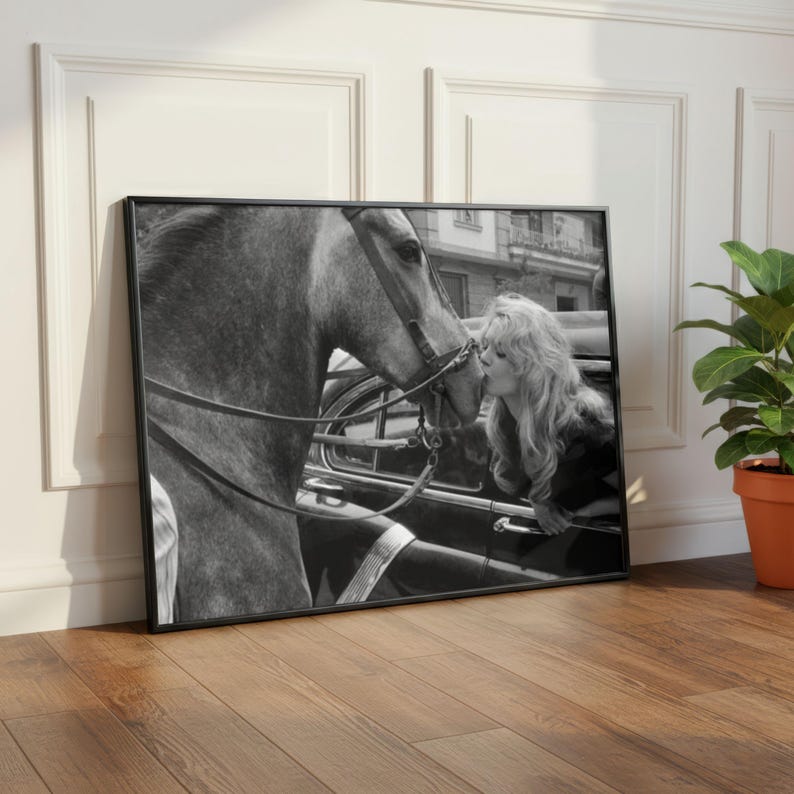 以下が含まれることがあります： 女性が馬にキスをしている白黒の額入りプリント。馬は手綱を着用しています。女性は長く波打つ髪をしており、車の窓から身を乗り出しています。プリントは木製の床に展示されています。