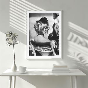 May include: Black and white framed print featuring a vintage photograph of Brigitte Bardot. She is seated in a director's chair with her name printed on the back. She wears sunglasses and a dark flower in her hair.