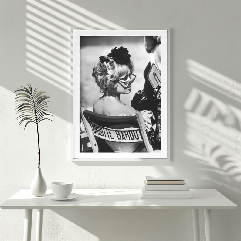 May include: Black and white framed print featuring a vintage photograph of Brigitte Bardot. She is seated in a director's chair with her name printed on the back. She wears sunglasses and a dark flower in her hair.