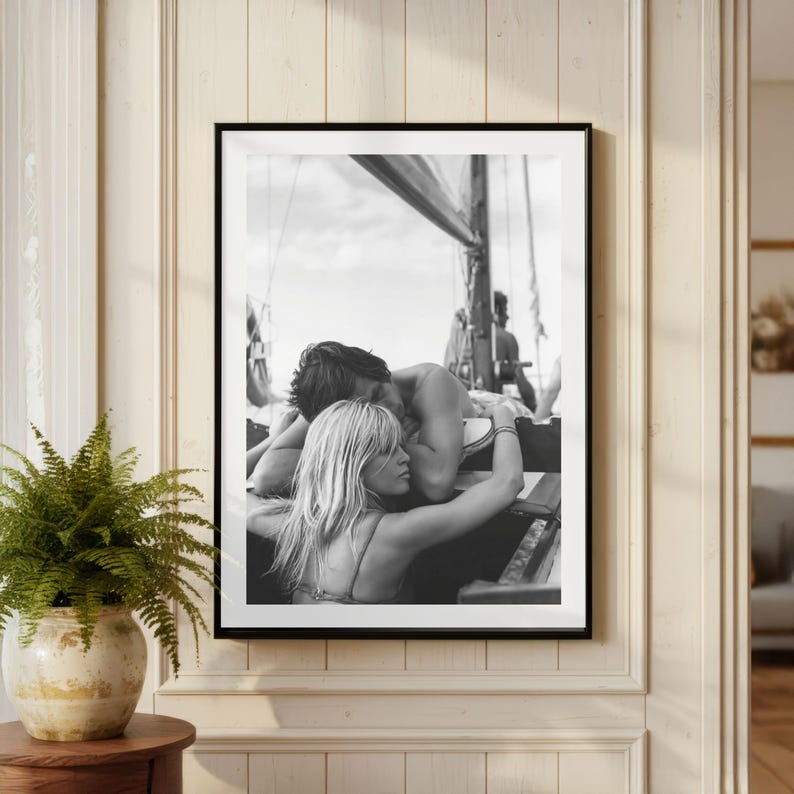 K&ouml;nnte beinhalten: Schwarz-Wei&szlig;-Druck in einem Rahmen, der ein Paar auf einem Segelboot zeigt, das sich umarmt. Das Bild zeigt einen Mann und eine Frau in inniger Umarmung, mit dem Bootsrigg und dem Himmel im Hintergrund. Eine romantische Szene.