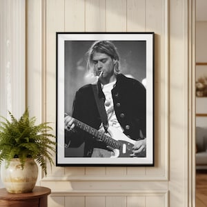Puede incluir: Impresión enmarcada en blanco y negro de un músico tocando una guitarra eléctrica blanca, con un cigarrillo en la boca. El músico lleva una chaqueta negra sobre una camiseta blanca. La impresión se muestra en una pared clara.