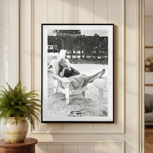 Puede incluir: Impresi&oacute;n enmarcada en blanco y negro de una mujer relaj&aacute;ndose en una silla de madera blanca. Lleva un top oscuro y una falda a cuadros. La impresi&oacute;n se muestra en una pared blanca, con un helecho en una maceta visible a la izquierda.