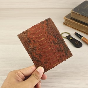 Könnte beinhalten: Ein rechteckiges Portemonnaie aus rotbraunem Schlangenhaut. Das Portemonnaie wird in einer Hand gehalten und zeigt das strukturierte Muster und die Farbvariationen. Ein Schlüsselanhänger, ein Stift und alte Bücher sind im Hintergrund zu sehen.