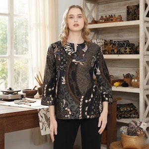 Op de afbeelding: Een zwarte blouse met een uniek batikpatroon in bruin, blauw en wit. De blouse heeft een uitgesneden halslijn en driekwartmouwen. Het model draagt een zwarte broek.