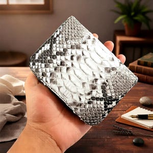 Op de afbeelding: Een bi-fold portemonnee met een slangenleerpatroon in tinten wit, grijs en zwart. De portemonnee is rechthoekig en lijkt van leer te zijn. De schubben zijn duidelijk zichtbaar, waardoor het een textuur heeft.