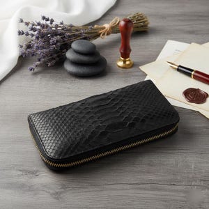 Puede incluir: Cartera de cuero negro con textura de piel de serpiente y cremallera dorada. La cartera rectangular est&aacute; cerrada. Otros elementos: lavanda, una pila de piedras, un sello de cera y un bol&iacute;grafo.