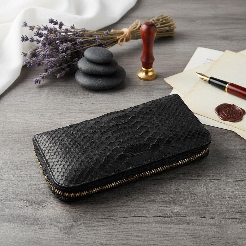 Puede incluir: Cartera de cuero negro con textura de piel de serpiente y cremallera dorada. La cartera rectangular est&aacute; cerrada. Otros elementos: lavanda, una pila de piedras, un sello de cera y un bol&iacute;grafo.