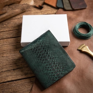 May include: A dark green bi-fold wallet featuring a textured snakeskin pattern. The wallet is positioned in front of a plain white rectangular box, showcasing a detailed scale-like surface finish across the exterior of the leather accessory.