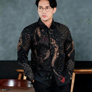 Op de afbeelding: Een zwart overhemd met een ingewikkeld bruin en rood batikpatroon. Het shirt met lange mouwen wordt gecombineerd met een zwarte broek. Het patroon van het shirt bevat bloemen en abstracte motieven.