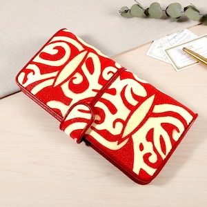 May include: A red and cream-coloured wallet featuring a distinctive swirling pattern. The wallet has a red leather trim and a matching strap closure. The design is striking, and the wallet appears to be made of high-quality materials.