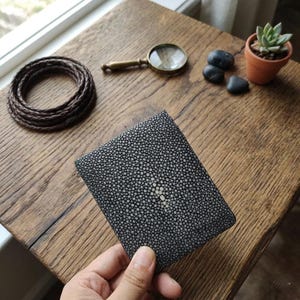 Peut inclure: Un portefeuille en cuir de raie noir et blanc avec un motif pointillé unique. Le portefeuille est tenu dans une main, mettant en valeur sa texture et son design. D'autres éléments sur la surface en bois comprennent une loupe, une plante succulente et des pierres décoratives.