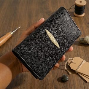 May include: Black leather wallet with a textured finish and a cream-coloured stingray design. The rectangular wallet is closed. The wallet is held in a person's hand.