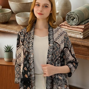 May include: An open-front jacket featuring a black, beige, and brown batik-style pattern with intricate floral and geometric motifs, three-quarter length sleeves, and a straight hem, worn over a plain white camisole and white trousers.