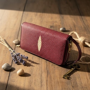 Könnte beinhalten: Eine weinrote Leder-Clutch mit strukturierter Oberfläche und einem cremefarbenen, ovalen Design. Die Clutch hat einen goldenen Reißverschluss und einen kleinen Griff. Das Bild befindet sich auf einer Holzoberfläche mit dekorativen Elementen.
