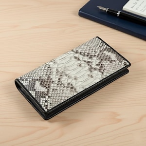May include: A rectangular wallet with a snakeskin pattern in shades of white, grey, and black. The wallet has a black border and appears to be made of leather. The wallet is closed and lying on a wooden surface.