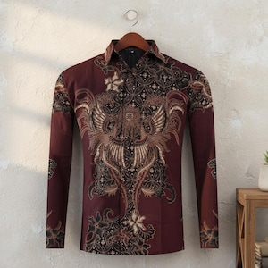Könnte beinhalten: Ein langärmeliges bordeauxfarbenes Hemd mit einem komplizierten Batikmuster. Das Design zeigt ein zentrales Vogelmotiv mit floralen und geometrischen Elementen in Braun- und Schwarztönen. Das Hemd wird auf einem Holzbügel präsentiert.