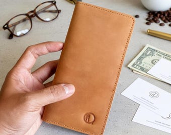 May include: A tan leather wallet with a stitched border and a debossed logo. The wallet is held in a hand, with eyeglasses, a pen, coffee, and money in the background. The wallet appears to be approximately 7 inches long.