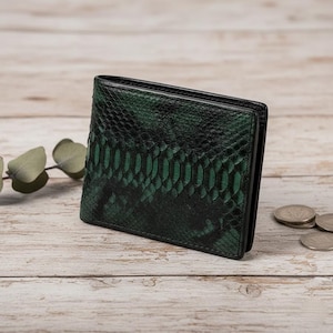 May include: A bi-fold wallet with a green snakeskin pattern. The wallet is made of leather and has a dark green and black color scheme. The wallet is sitting on a light wood surface.