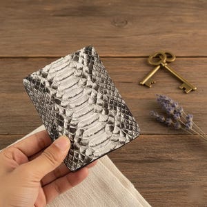 Può includere: Un portafoglio a due ante con un motivo a pelle di serpente nei toni del grigio e del bianco. Il portafoglio è tenuto in mano da una persona, con una superficie di legno sullo sfondo. Sono visibili anche un paio di chiavi antiche e rametti di lavanda.