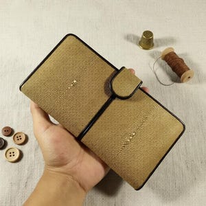 Könnte beinhalten: Eine hellbraune Ledergeldbörse mit strukturierter Oberfläche und schwarzem Rand. Die Geldbörse ist geöffnet und zeigt das Innere. Ein Fingerhut, eine Garnrolle und Knöpfe befinden sich im Hintergrund.