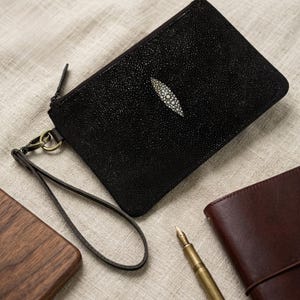 Puede incluir: Clutch de cuero negro con un distintivo diseño de raya y un diseño ovalado blanco. El clutch tiene cierre de cremallera, una correa de muñeca y un cierre dorado. Otros elementos en la imagen incluyen un bloque de madera, un bolígrafo y un cuaderno de cuero marrón.