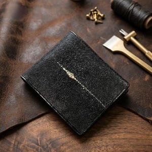May include: Black leather wallet with a stingray skin detail. The wallet is rectangular and features a white stripe. It rests on a brown leather surface, with tools and supplies visible in the background.
